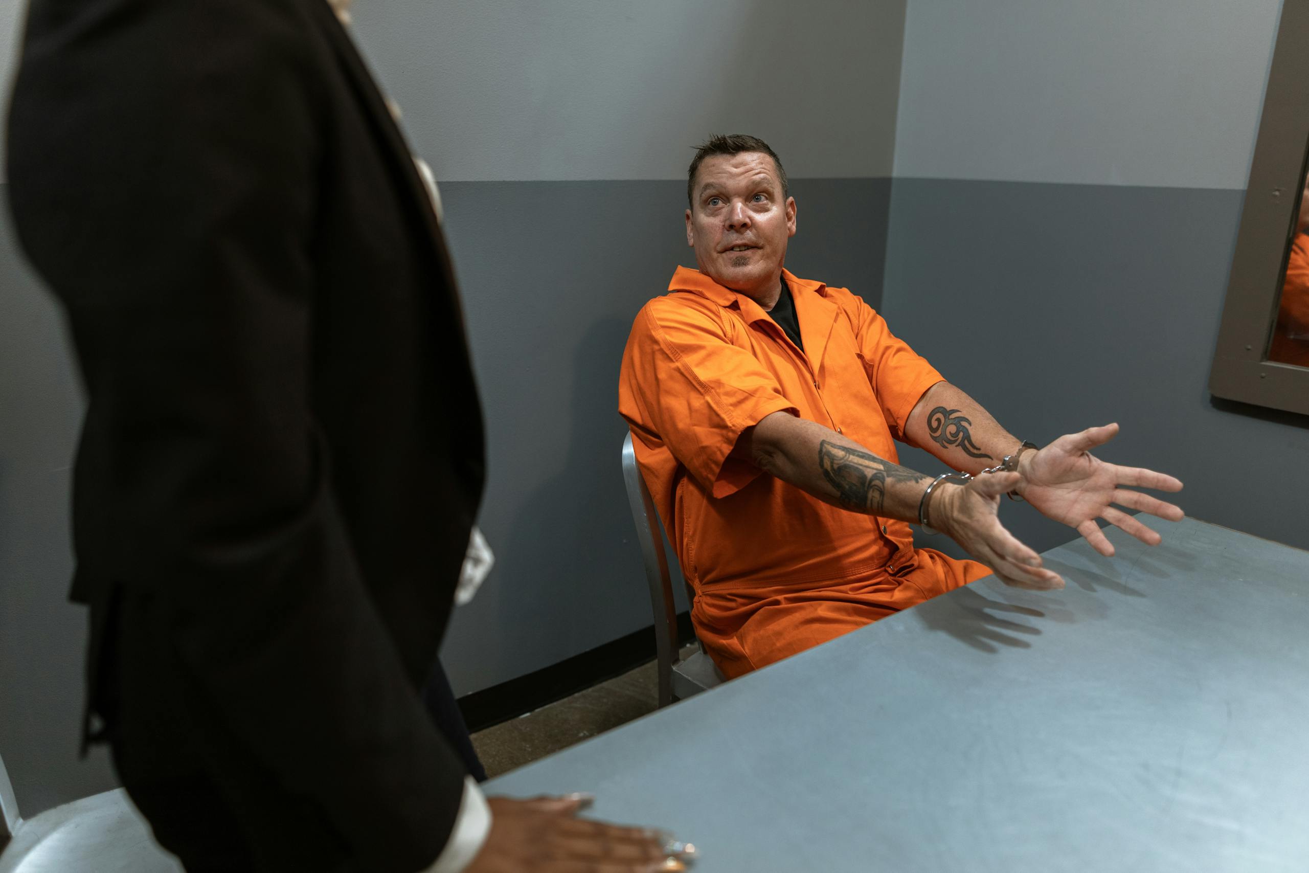 A prisoner in an orange jumpsuit sits handcuffed at a table in an interrogation room.