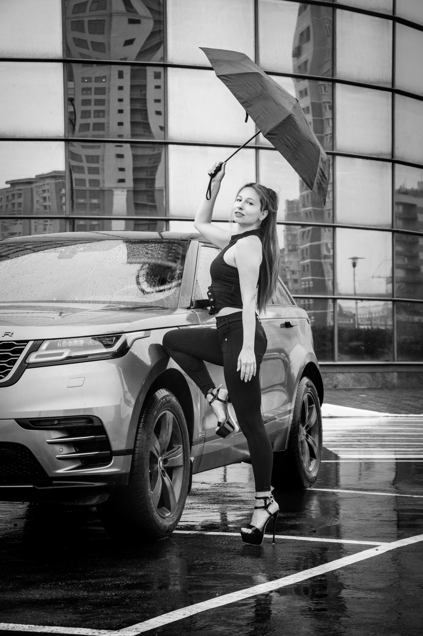 Black and white urban portrait of a fashionable woman in heels posing with an umbrella beside a luxury car.