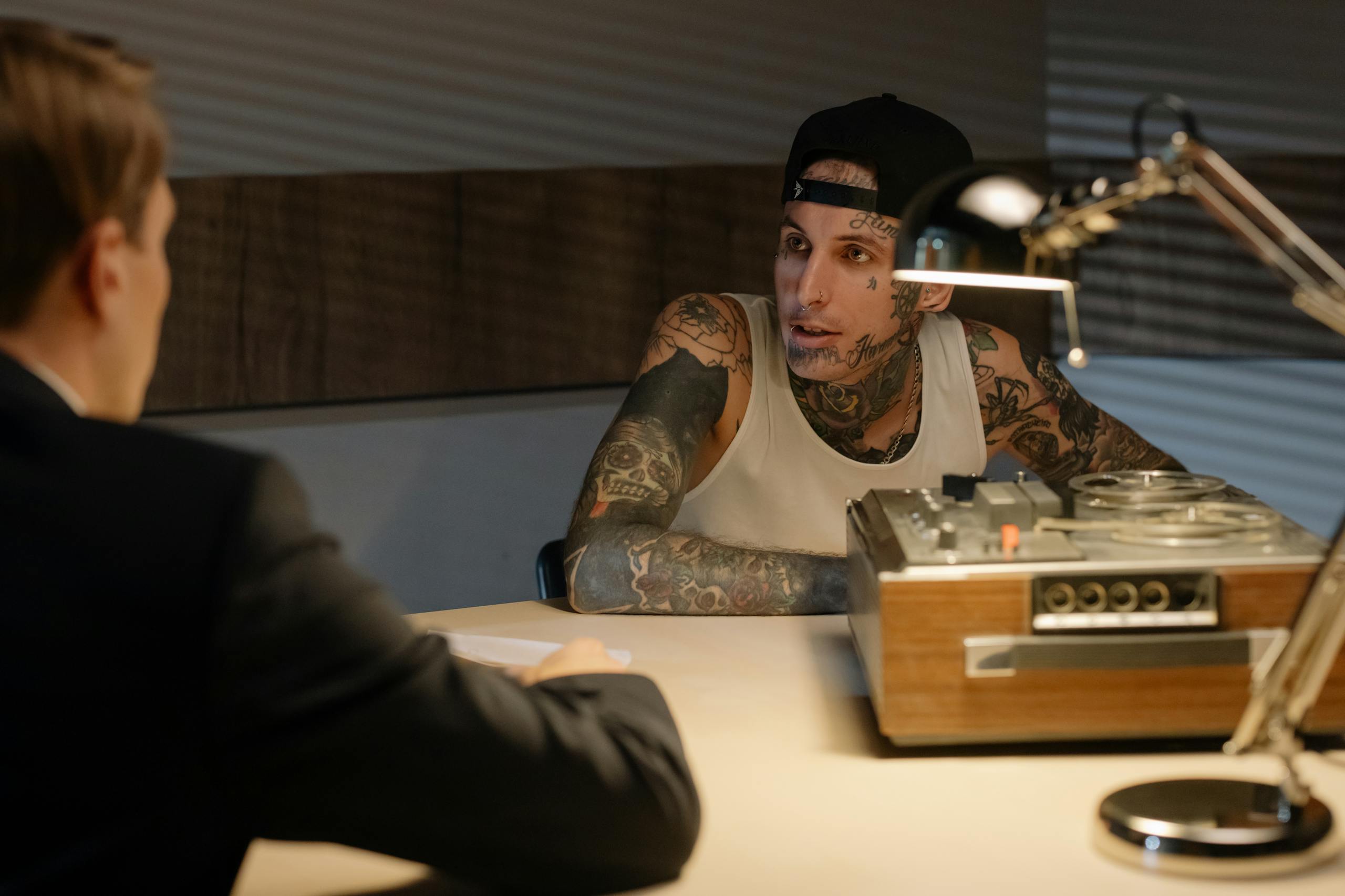 Tattooed man being questioned in an interrogation room, under warm lighting.